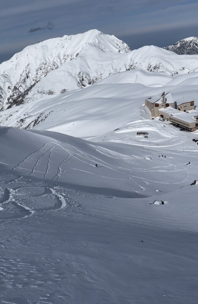 立山の室堂エリアで広大なオープンバーンを滑走するスノーボーダーと残されたトラック跡、一番奥に見えるのは大日三山