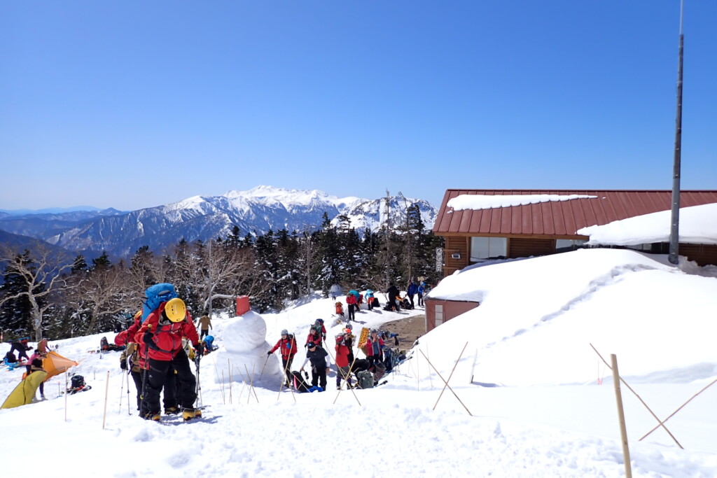 冬の西穂山荘前の様子と登山者、奥には乗鞍岳の雪景色